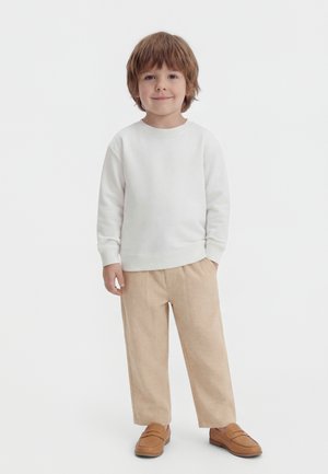 Mladý chlapec stojaci s rukami v vreckách, oblečený v bielom tričku s dlhým rukávom, béžových nohaviciach a hnedých mokasínach na bielom pozadí.