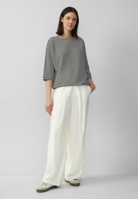Zwart-witte gestreepte top met driekwartmouwen, gecombineerd met losse, crèmewitte broek en lichte sneakers, staand tegen een neutrale achtergrond.