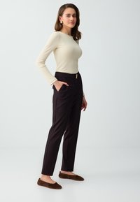 Femme portant un haut crème à manches longues, un pantalon noir taille haute et des ballerines marron, debout avec une main dans la poche sur fond uni.