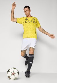 Yellow soccer jersey with a textured pattern, black accents, and logo, paired with white shorts and black socks with stripes. Black soccer ball nearby.