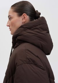 Giacca a cappuccio marrone con un'imbottitura trapuntata, dotata di un colletto alto e di cordini regolabili. Il materiale sembra morbido e leggero.