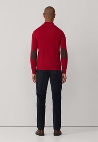 Roter Strickpullover mit braunen Ellenbogenpatches, geripptem Kragen und Bündchen. Getragen mit dunklen Hosen und braunen Schuhen, von hinten gesehen.