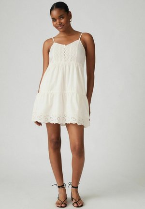 Witte zonnejurk met spaghettibandjes, geborduurd lijfje, gelaagd ontwerp, gegolfde zoom en uitgesneden bloemenpatronen. Draagt met zwarte sandalen.