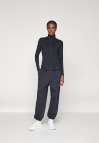 Schwarze Zip-Jacke mit hohem Kragen, kombiniert mit dunkelgrauen, schmal zulaufenden Jogginghosen und weißen Sneakers. Glattes Material mit minimalistischen Marken-Details.