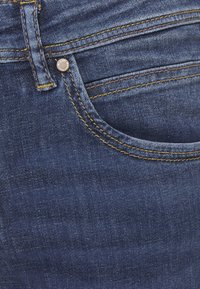 Mörkblå denimjeans med kontrasterande gula stygn, klassisk femficksdesign och en metallknappstängning framtill.