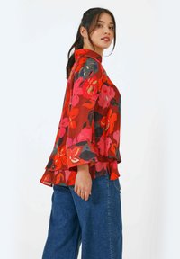 Rode bloemenblouse met klokmouwen, met levendige roze en donkergroene bloemen. Lichtgewicht, vloeiende stof valt losjes, met een gelaagde zoom.