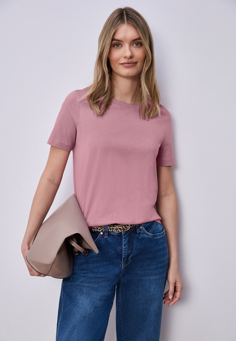Femme aux cheveux blonds portant un t-shirt violet clair, un jean bleu avec une ceinture à imprimé léopard, tenant un sac à main beige sur un fond uni.