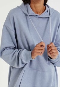 Sweat à capuche ample bleu clair avec poche kangourou, capuche à cordon, poignets côtelés et détails cousus. La texture du tissu doux est visible.