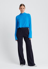 Heldere blauwe lange mouw top met hoge hals, gecombineerd met donkere wijde broekspijpen. De outfit heeft een textuurstof met een gladde afwerking.