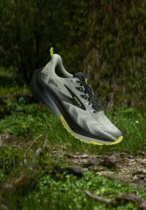 Světle šedá a černá Brooks běžecká bota s neonově žlutými akcenty plovoucí nad kamenitou travnatou stezkou.