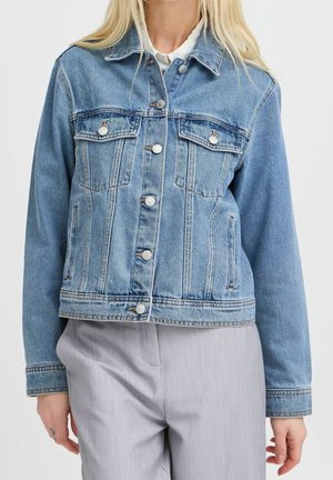 Žena nosící světle modrou džínovou bundu se zapínáním na knoflíky a kapsami na prsou a světlé šedé kalhoty, stojící před jednobarevným pozadím.