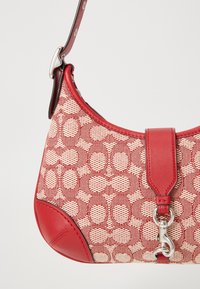 Sac à main rouge et beige à motifs avec garniture en cuir, fermoir argenté et bandoulière réglable sur fond uni.