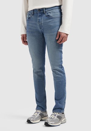 Slim-fit blue jeans worn with silver and gray sneakers and a white long-sleeve top, standing against a plain light background.