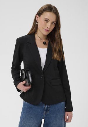 Frau mit langen braunen Haaren, die ein schwarzes Blazer über einem weißen Oberteil, blaue Jeans trägt, eine schwarze Clutch hält und Perlenohrringe sowie eine schwarze Halskette trägt.
