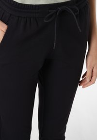 Zwarte joggingsbroek met elastische tailleband en trekkoord. Voorzien van zijzakken en een gladde, elastische stoftextuur.