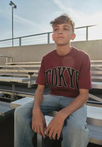 Teenager-Junge trägt ein Tokyo-T-Shirt und Jeans, sitzt auf Metalltribünen im Freien mit Sonnenlicht und Himmel im Hintergrund.