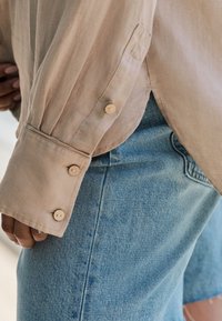 Light beige, long-sleeve shirt with buttoned cuffs, partially tucked into light blue denim jeans with a faded texture and subtle stitching details.