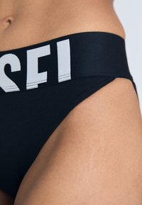 Zwarte bikini onderkant met een brede elastische tailleband en contrasterende witte logotekst. Glad materiaal met een aansluitende snit en laag uitgesneden ontwerp.