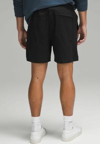 Shorts noirs avec une taille élastique, une poche arrière et une coupe décontractée. Le modèle porte des baskets blanches et des chaussettes, vu de dos.