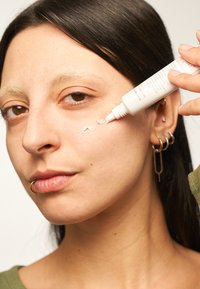 Dispensador de tubo blanco aplicado sobre la piel, con una textura suave y cremosa; el modelo tiene el cabello castaño claro, maquillaje mínimo y piercing de oro.