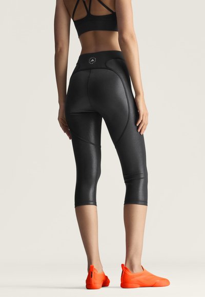 Kvinde iført skinnende sorte cropped leggings, en sort sports-bh og lyse orange slip-on atletik sko, stående med ryggen mod fronten.