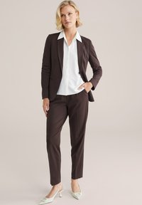 Hnedý oblek na mieru so bielou košeľou. Sako má ostré klopy; nohavice sú priliehavé. Výrazné mentolové lodičky dotvárajú outfit.