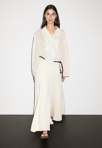 Cardigan beige léger sur une chemise blanche ; jupe fluide crème avec une ceinture en perles noires ; sandales roses ; texture lisse et design simple.