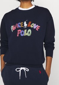 Námořnický modrý svetr s kulatým výstřihem. Vícebarevný text "PEACE LOVE POLO" a bílý logo polistova hráče na přední straně. Měkká bavlněná tkanina.