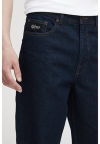 Pantalones vaqueros de mezclilla azul oscuro con corte recto, costuras naranjas y un pequeño logo en el bolsillo de la cintura. La textura es suave y resistente.