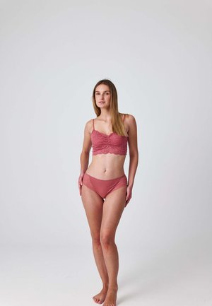 Femme debout pieds nus sur un fond uni, portant un bralette en dentelle rose poussiére assorti et des sous-vêtements, regardant légèrement sur le côté.