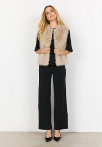 Faux-Fur-Weste in hellbeige mit einer strukturierten, plüschigen Oberfläche, getragen über einem schwarzen Metalltop und weiten schwarzen Hosen, kombiniert mit schwarzen Absätzen.