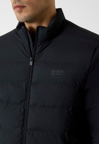 Schwarze Steppjacke mit hohem Kragen, Reißverschluss und "GUESS TECH STRETCH"-Logo. Glatte Textur und gesteppte Musterung.
