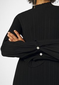 Persona che indossa un top a maniche lunghe a coste nero con polsini bottonati, braccia incrociate e un orecchino d'oro pendente visibile.