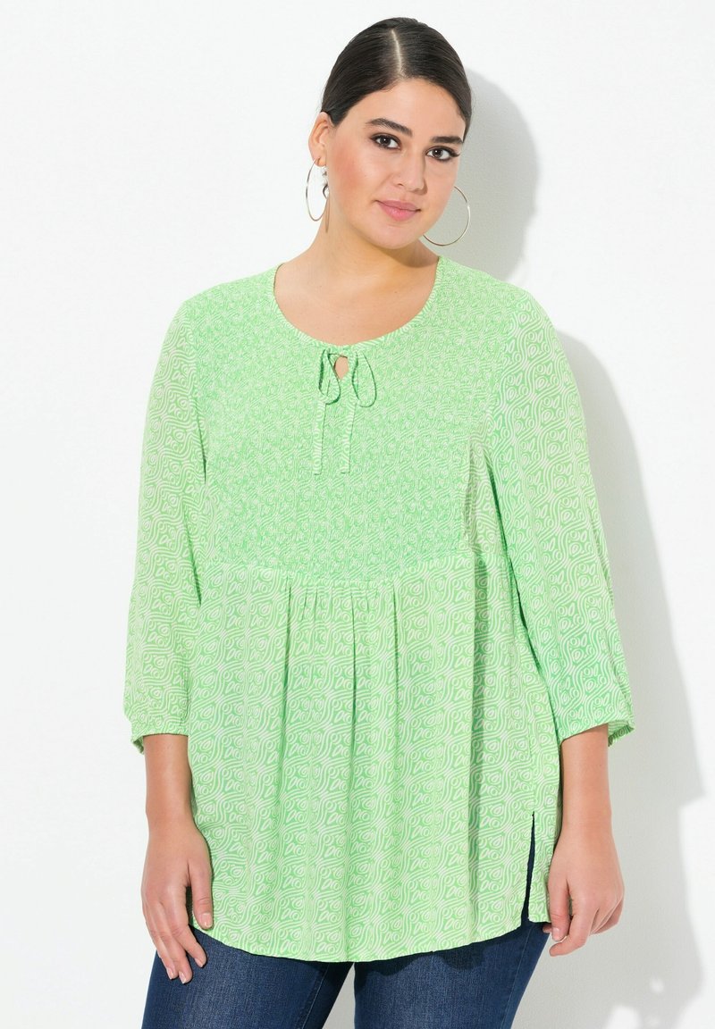 Blouse verte à motif avec des manches trois-quarts, détail en trou de serrure à l'avant, ourlet arrondi et fentes latérales. Texture de tissu douce, coupe décontractée.