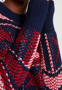 Persoon draagt een marineblauwe, rode en witte gebreide trui met ribbels aan de manchetten, terwijl hij met één hand gedeeltelijk zijn gezicht bedekt.