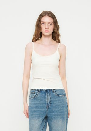 Ung kvinne med bølgete hår iført en hvit, ermeløs topp med blonder og blå denimjeans, stående foran en ensfarget hvit bakgrunn.