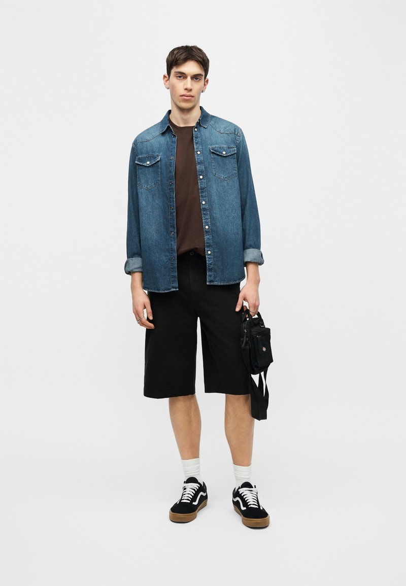 Jeune homme debout portant une chemise en jean bleu par-dessus un t-shirt marron, un short noir jusqu'aux genoux, des baskets noires, et tenant un petit sac noir.