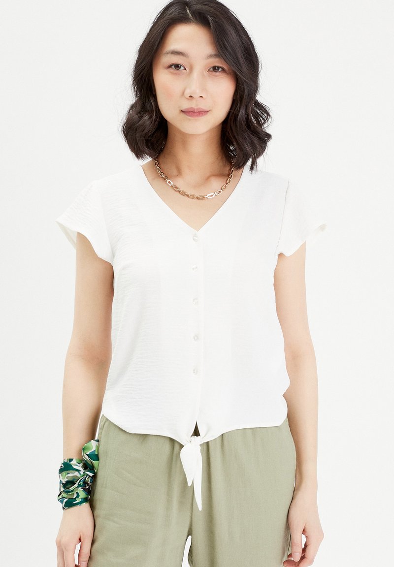 Blouse blanche à manches courtes avec un décolleté en V, boutons sur le devant et un détail à nouer en bas. Portée avec un pantalon vert et un chouchou à motifs.