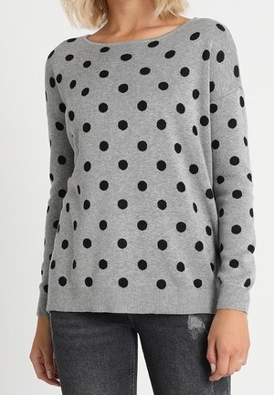 Woman wearing a gray long-sleeve sweater with black polka dots and dark gray distressed jeans, standing with arms relaxed by sides.