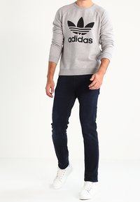 Gray sweatshirt with black Adidas logo, round neckline, ribbed cuffs, paired with dark blue jeans and white sneakers. Simple design, casual fit.