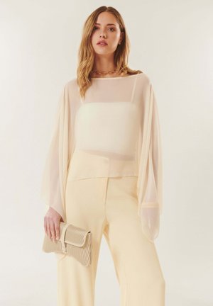 Femme portant un chemisier crème transparent sur un caraco et un pantalon crème, tenant une pochette beige texturée, debout devant un fond clair.