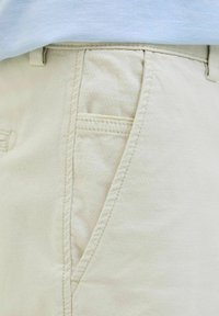 Gros plan sur la poche d’un pantalon beige avec des détails de couture visibles et un haut en tissu blanc partiellement rentré.