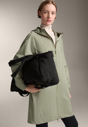 Helles grünes Zip-Jackett mit Kapuze und seitlichen Taschen, gehalten über einer schwarzen, gepolsterten Tasche mit Riemen. Glattes Material und lässiges Design.
