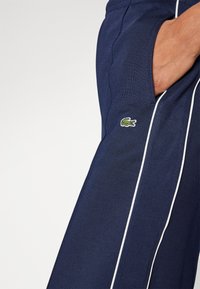 Navy blue pants with a textured fabric, featuring white side stripes and a green embroidered crocodile logo on the pocket.