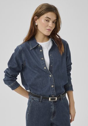 Mladá žena oblečená v tmavomodrej rifľovej košeli na gombíky cez bielu blúzku, tmavomodré rifľové nohavice, čierny opasok a strieborné náušnice, pózujúca s jednou rukou za chrbtom.