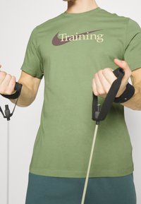Camiseta verde oliva con el texto "Entrenamiento" en color beige y púrpura oscuro. Persona sosteniendo bandas de resistencia con empuñaduras acolchadas negras.