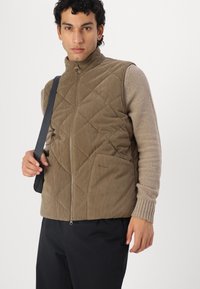 Braune, gesteppten Cord-Veste mit hohem Kragen, Frontreißverschluss und zwei aufgesetzten Taschen. Ärmel aus strukturiertem beigem Stoff. Schwarze Hose.