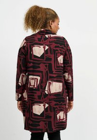 Pitkä musta takki, jossa on geometrinen kuvio burgundinpunaisena ja beigenä, rento istuvuus ja kolmanneksen mittaiset hihat, valmistettu joustavasta kankaasta.