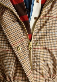 Jachetă cu fermoar cu pattern în carouri, în nuanțe de brun, bej și albastru. Dispune de un fermoar și capse aurii, cu un tricou stratificat dedesubt care evidențiază tonuri de roșu și brun.