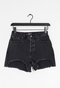 Schwarze Jeansshorts, die an einem Kleiderbügel hängen. Besitzen einen ausgefransten Saum, fünf Taschen und kupferfarbene Hardware-Details.
